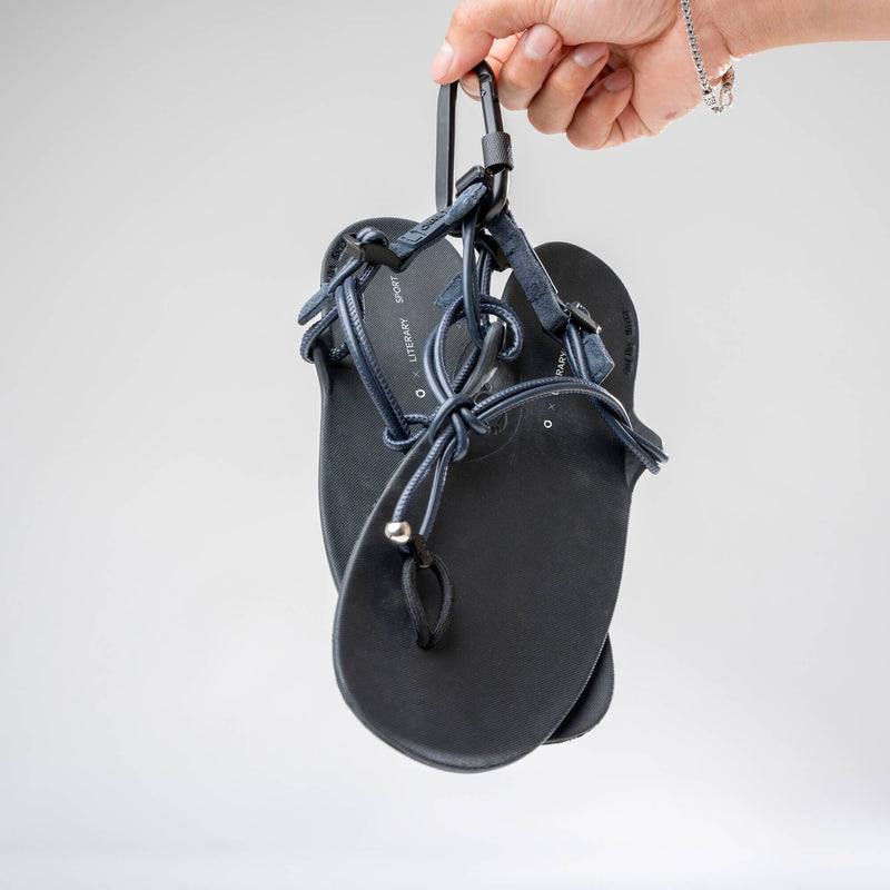 Xero Shoes Literary Sport Genesis Navy Strap Black Sandal with a Hand Holding the Pair of Sandals by a Carabiner